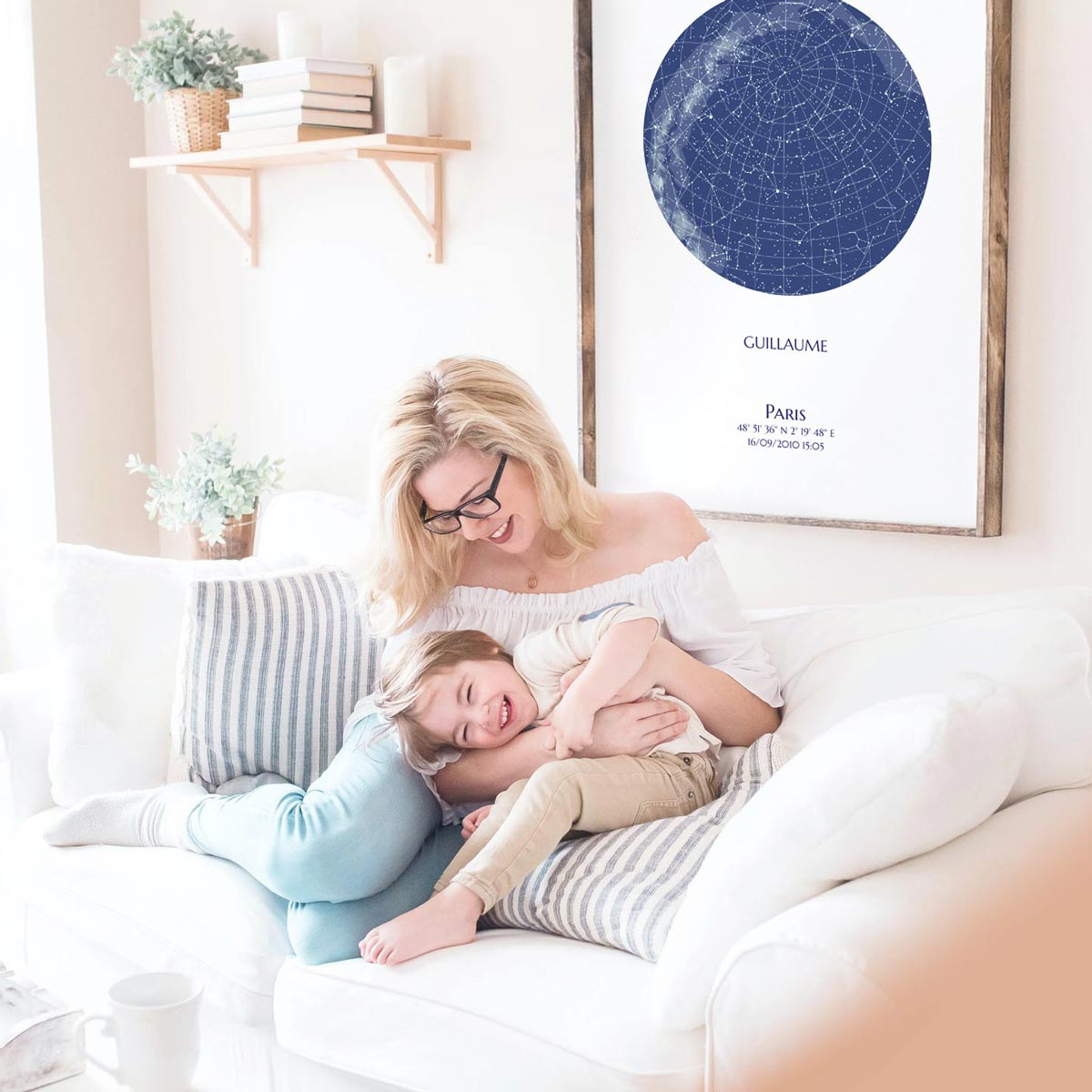 Une carte du ciel personnalisée de couleur bleu est accrochée au mur d'un salon où jouent et rigolent une maman avec son fils sur un canapé